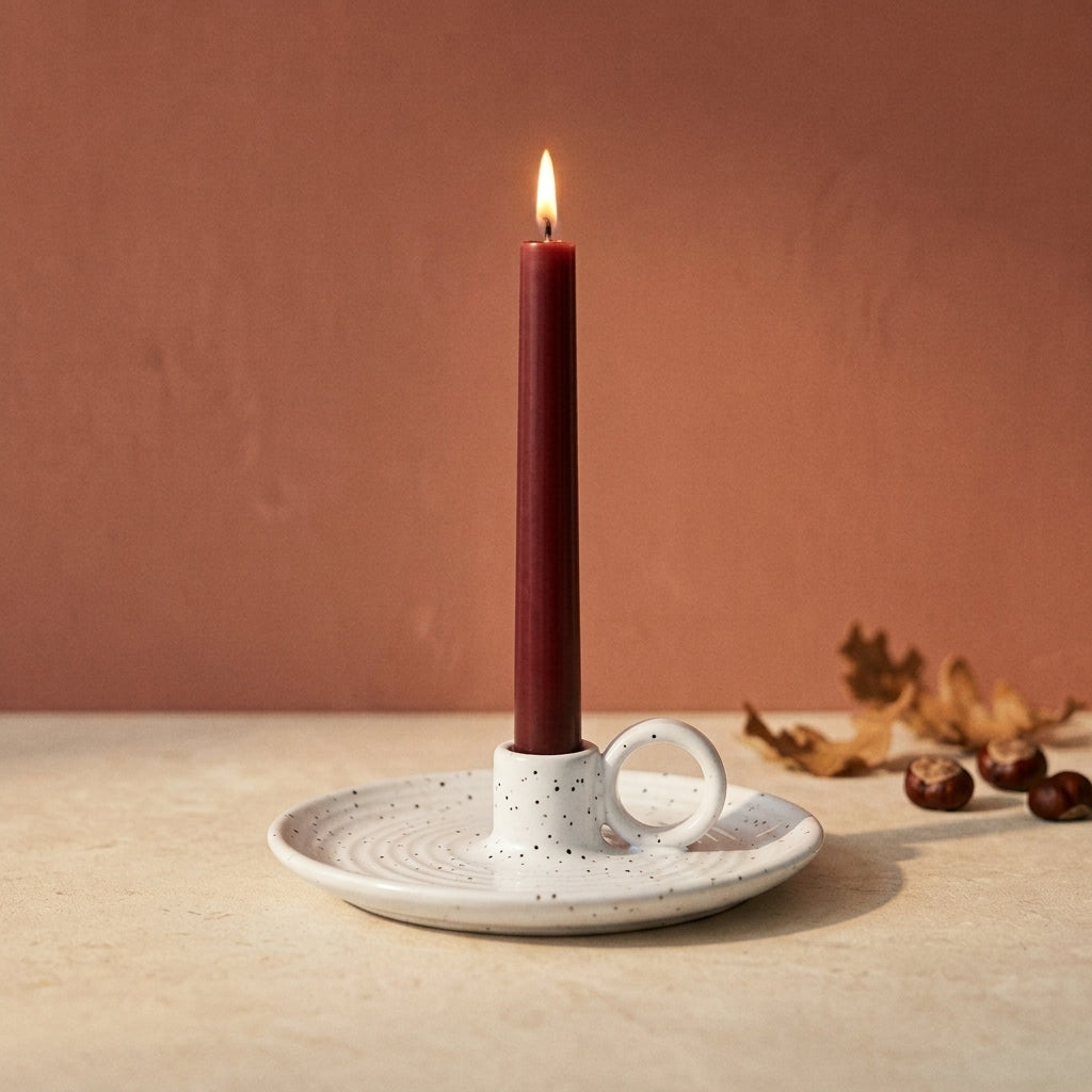 Speckled white ceramic chamberstick holding a lit burgundy taper candle, with conkers and autumn leaves